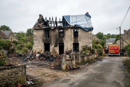 Maison de village incendiée, charpente effondrée et bâche bleue sur le toit, gravats devant la façade
